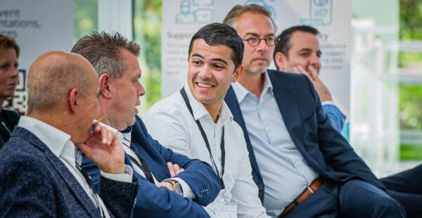 A group of five people sit side by side in a professional setting, engaged in an empowering conversation. One man in the center, wearing a white shirt, is smiling and looking at the man next to him. The others are listening attentively. Business attire and conference badges are visible.