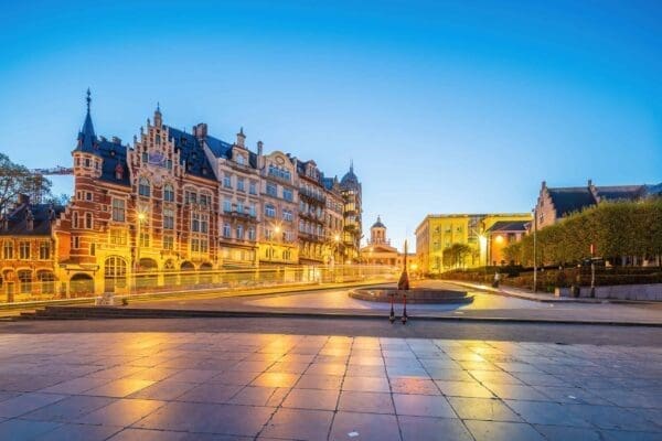 A twilight scene of Mont des Arts in Brussels, Belgium, showcasing historic, ornate buildings illuminated with warm lights. A lone figure stands near the circular fountain in the center, perhaps reflecting on the transition to e-Invoicing. The sky is clear with a deepening blue hue.