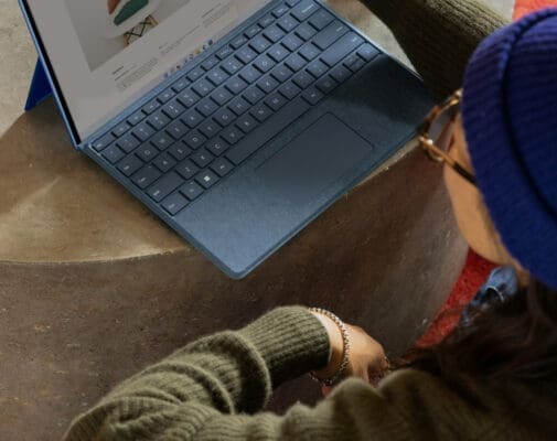 A person wearing a blue beanie and green sweater uses a laptop on a round concrete surface, reviewing their portfolio while searching for important SEO keywords on a product page displayed on the screen.